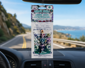 Package of Sugar Skull Smoke Odor Exterminator hanging from a car's rearview mirror with a scenic road and ocean view in the background.